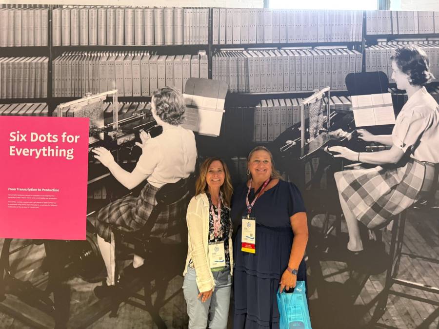 Cindy stands with a colleague from South Dakota in front of a large image of workers producing braille textbooks at the Dot Experience Museum in Louisville, KY.